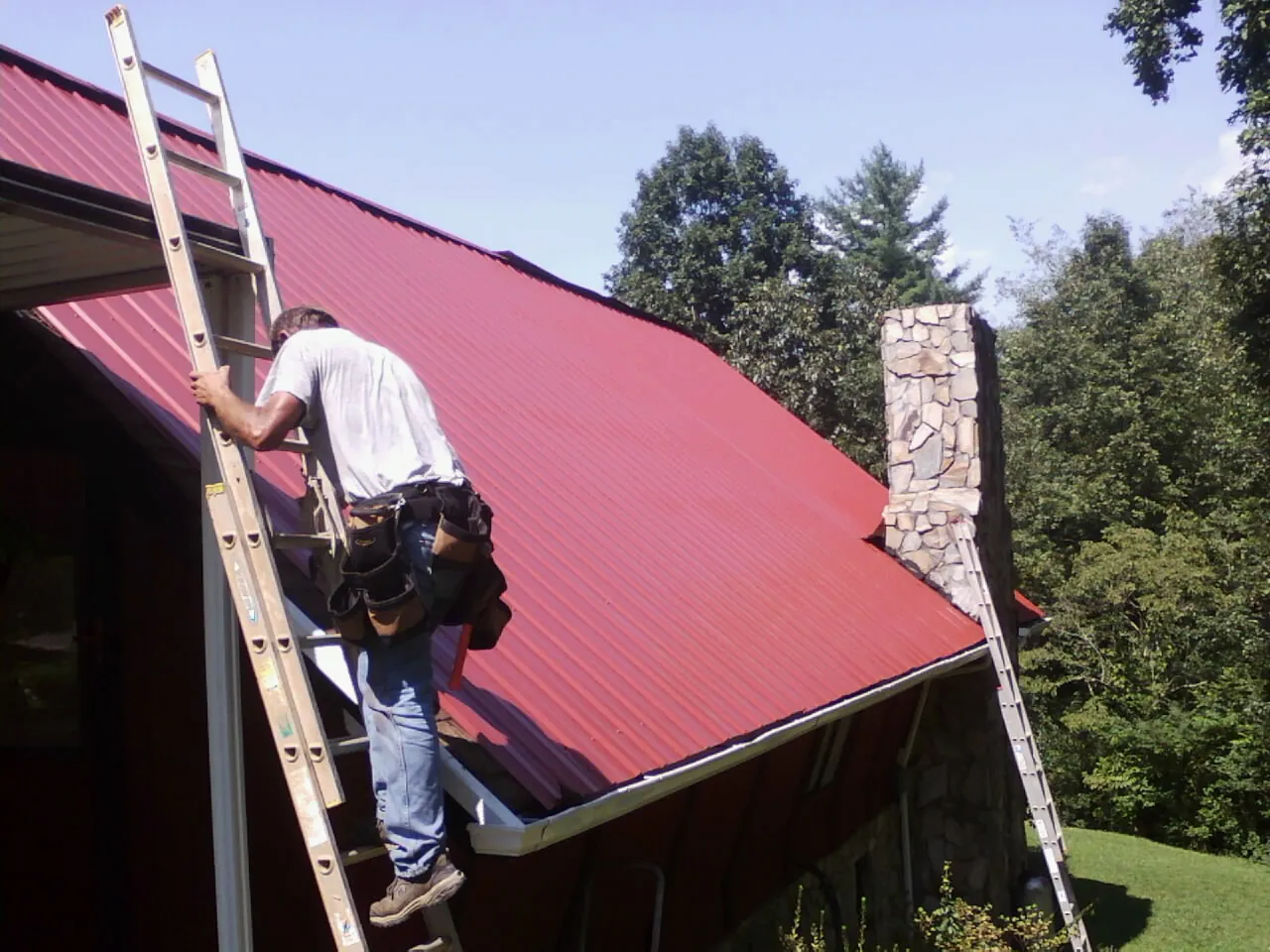 Roofer on a ladder beside a red metal roof and stone chimney — Soffit & Fascia Repair by Roofing Brothers in Glasgow
